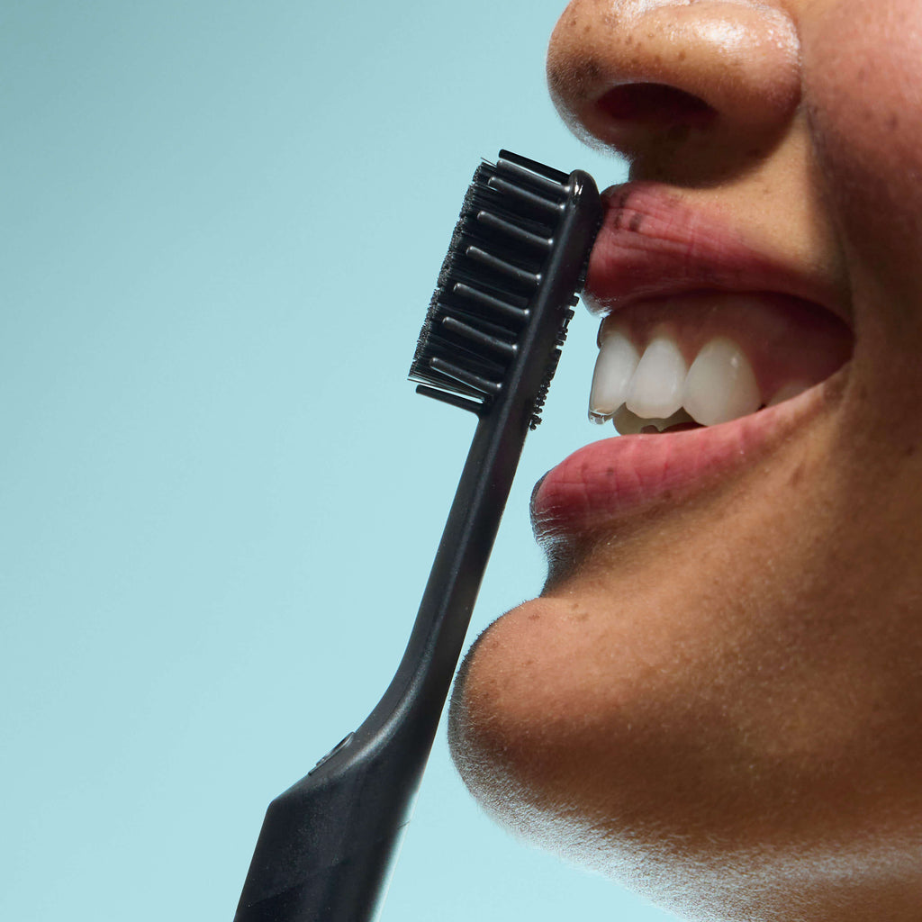 Close-up of a person smiling as a black Sonic toothbrush approaches their teeth, against a light blue background.